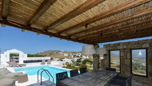 An outdoor pool area with a wooden pergola, a dining table with chairs, and a view of a coastal village. - ISholidays Mykonos villa Pearl (Ornos)