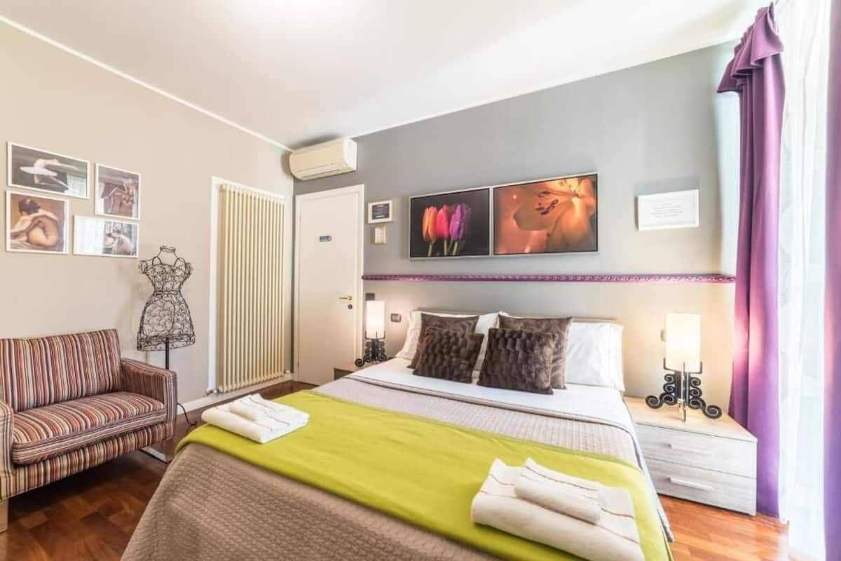 A hotel room with a bed, a sofa, a lamp, and artwork on the wall.
