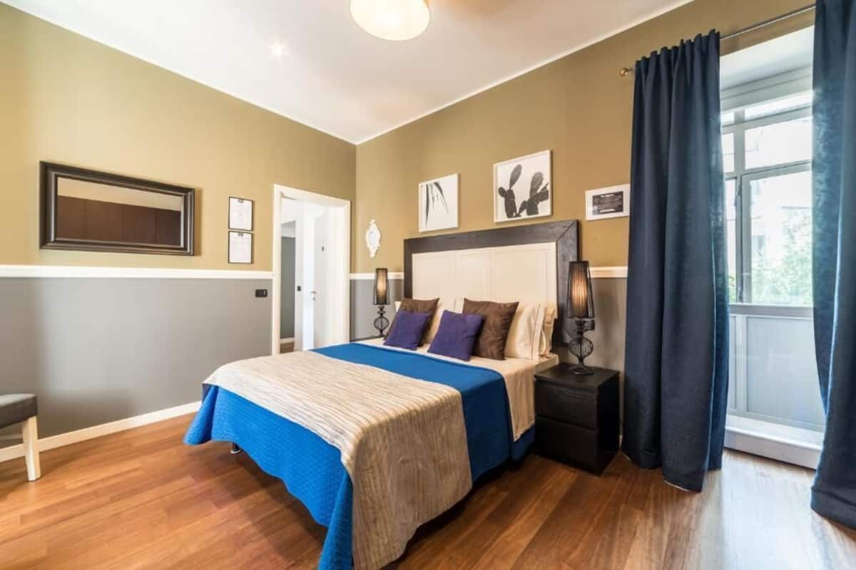 A bedroom with a bed, bedside tables, a nightstand, a window with curtains, and wall art.