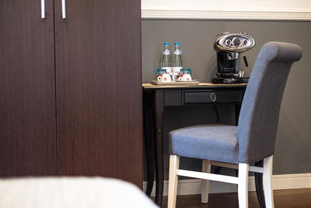 A hotel room with a dark wood wardrobe, a desk with a coffee machine, and a grey upholstered chair.