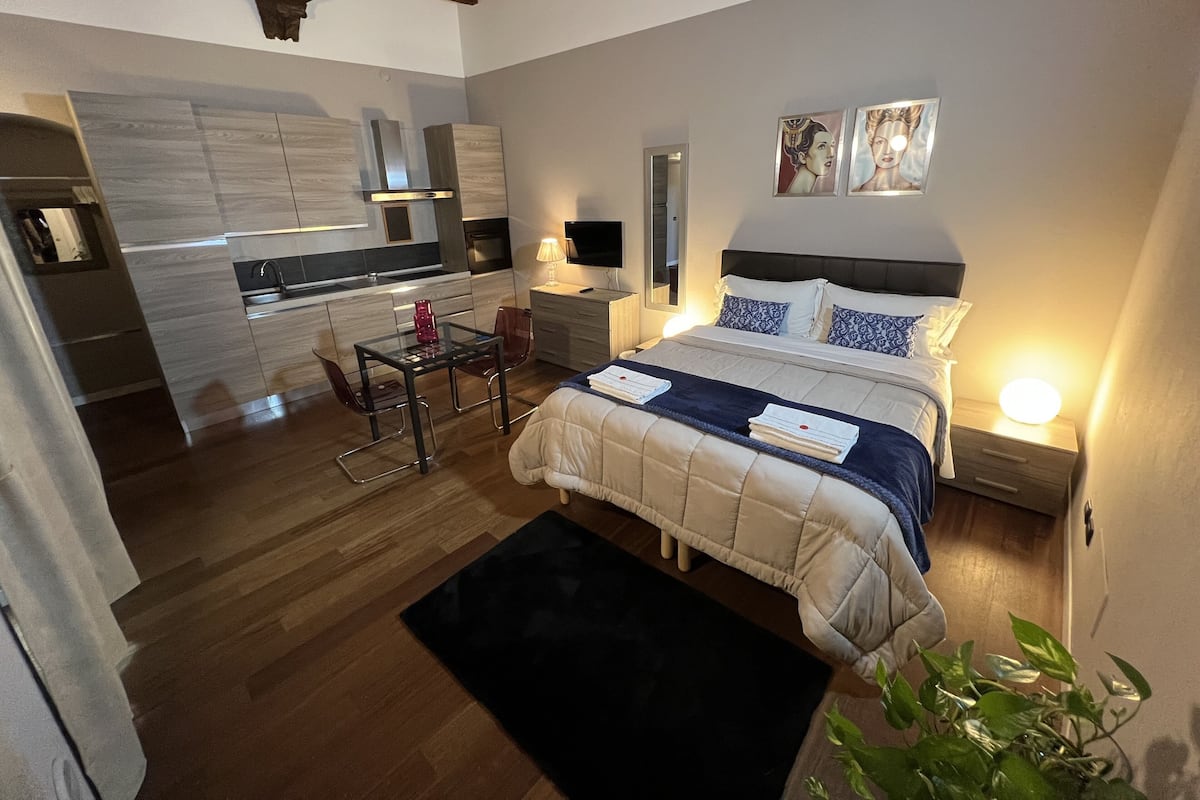 A modern bedroom with a large bed, bedside tables, a desk, and a chair. There is a kitchen area with a dining table and chairs.