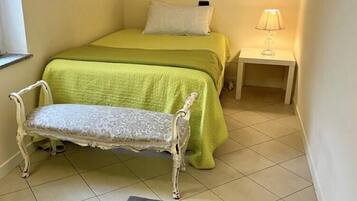 A small, neatly arranged bedroom with a bed, bench, bedside table, lamp, and a floral artwork on the wall.