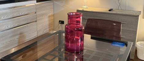 A modern kitchen with a glass-top dining table, a pink vase, a TV, and wooden cabinetry.