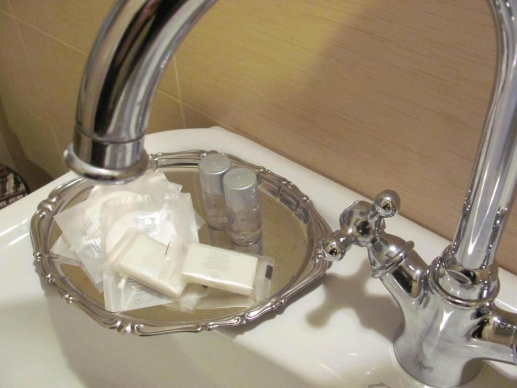 Ein Badezimmerwaschbecken mit chromfarbenem Wasserhahn, einer klaren Glasbecken und Badaccessoires, darunter eine Seife und zwei Flaschen.