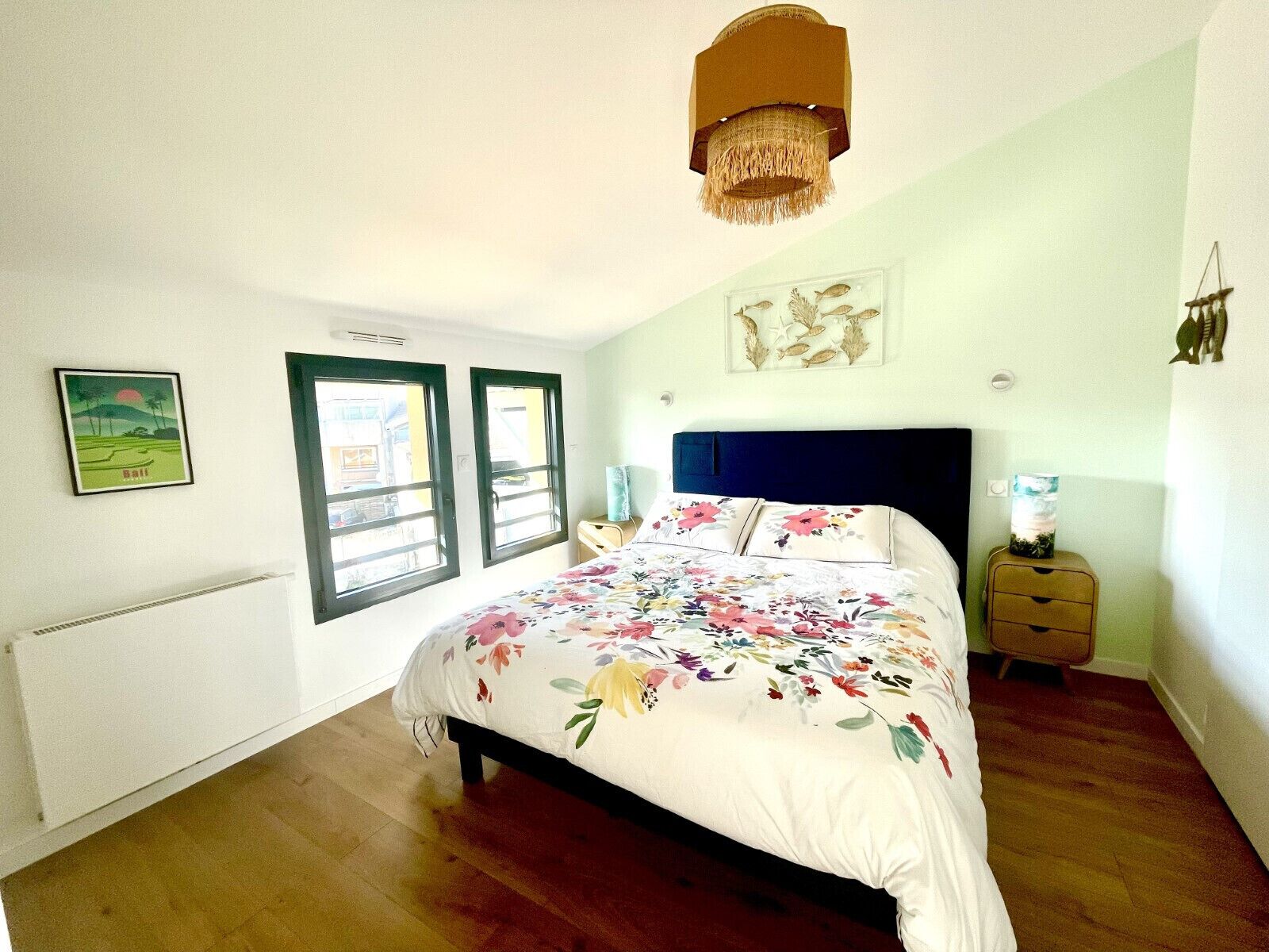 A bedroom with a bed, two windows, a radiator, a nightstand, and a hanging light fixture.