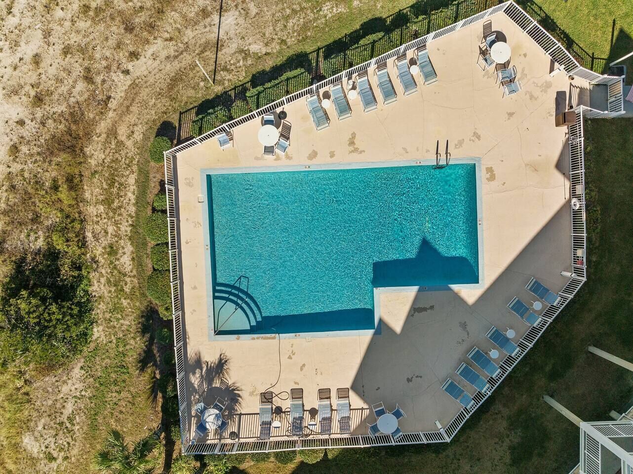 An aerial view of a rectangular swimming pool with lounge chairs and sunbeds around it, surrounded by a fence.