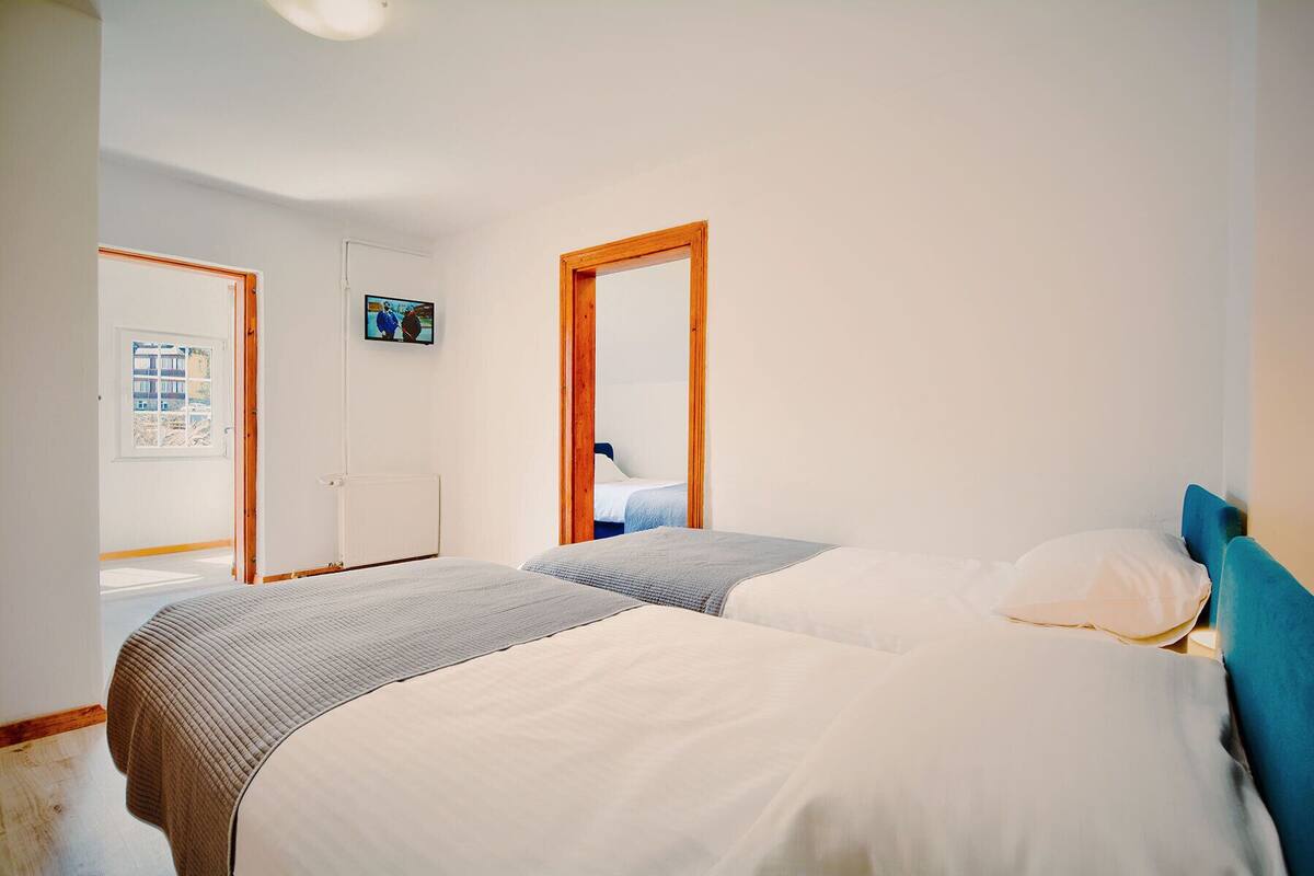 A bedroom with two beds, a television on the wall, and a window with a view of buildings.