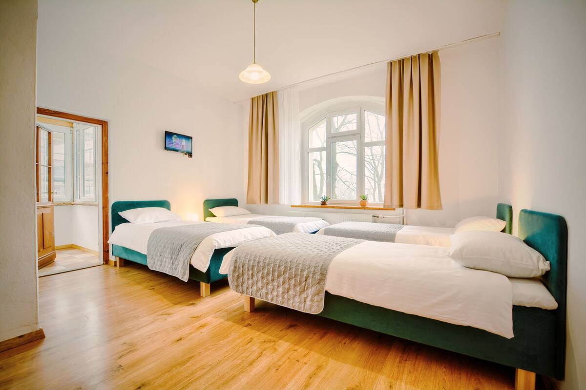 A room with three single beds, wooden flooring, a large window with curtains, and a small television on the wall.