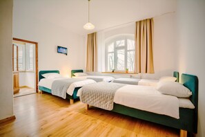 Een kamer met drie enkele bedden, houten vloeren, een groot raam met gordijnen en een kleine televisie aan de muur.