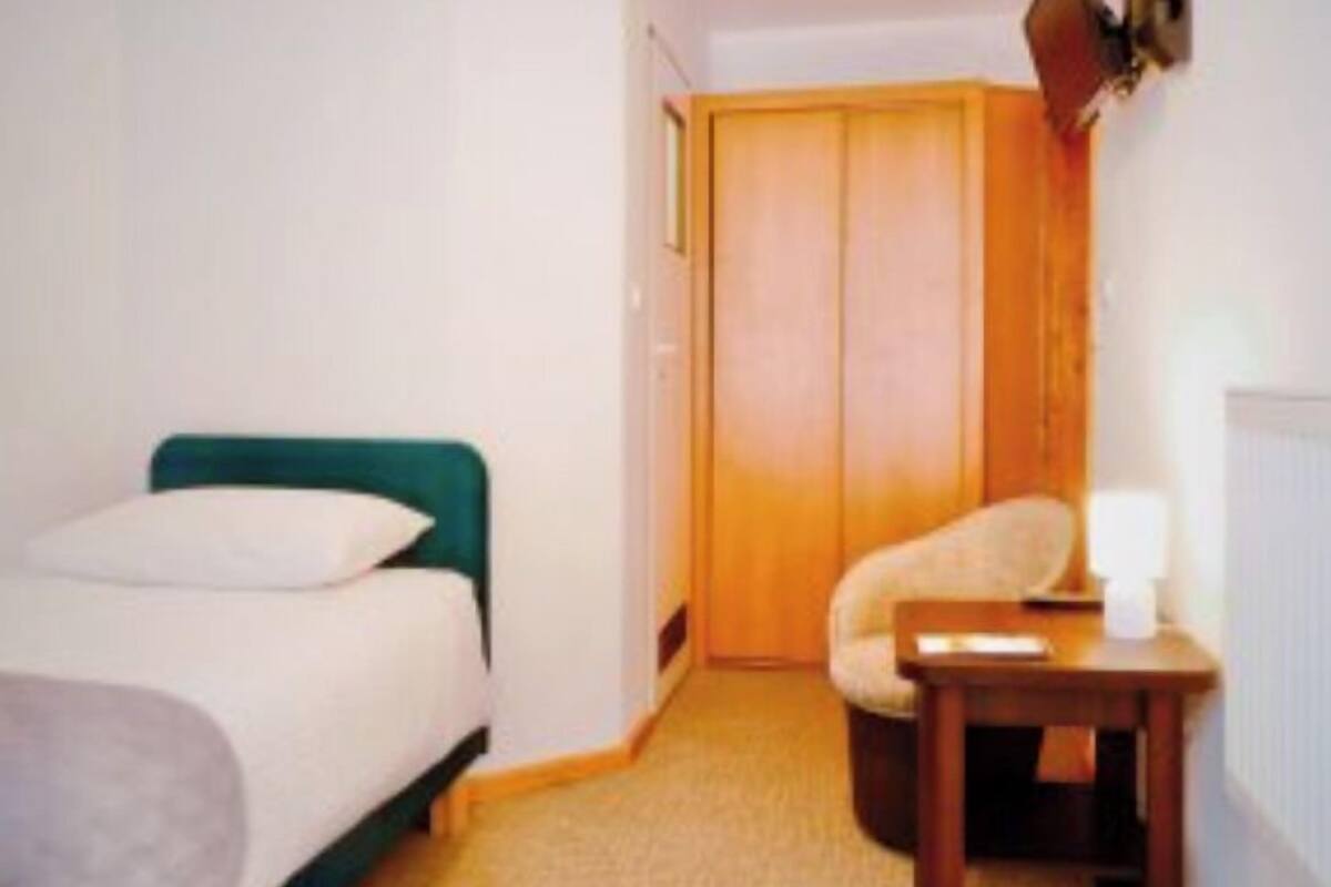 A small, single-bed room with a green headboard, a wooden wardrobe, a small table with a lamp, and a chair.