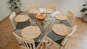 A round glass table with six wooden chairs, each with a woven placemat. A terracotta bowl with decorative items is placed in the center.