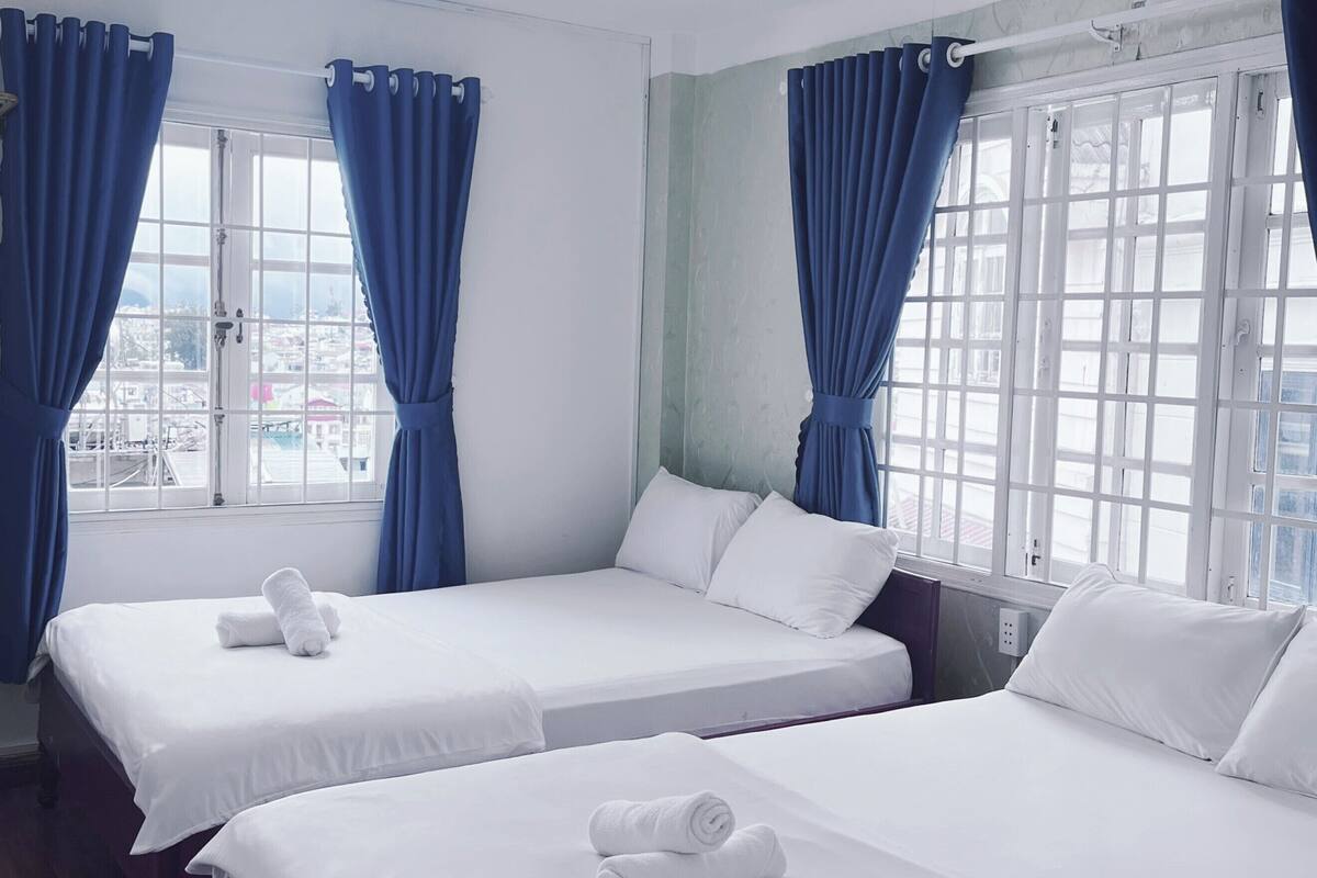 A hotel room with two beds, white bedding, blue curtains, and a view of the city through large windows.
