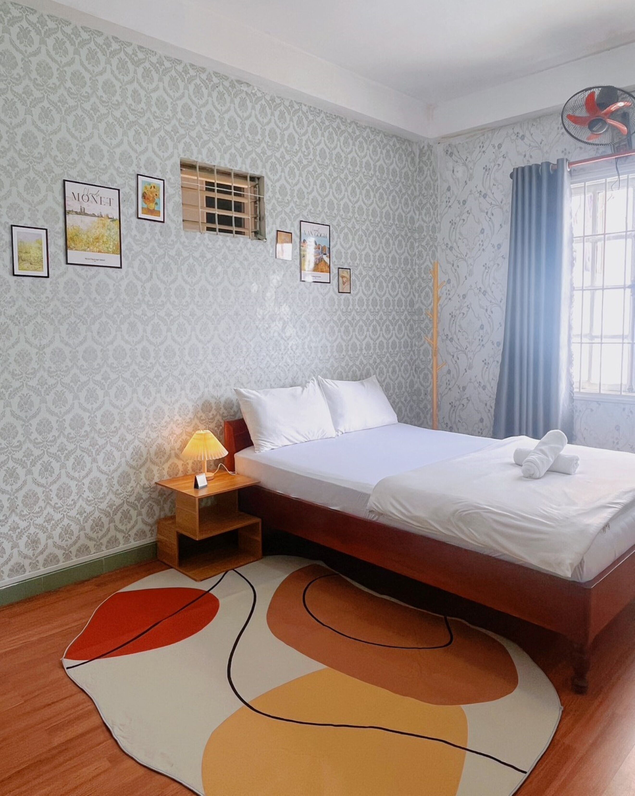 A neatly arranged bedroom with a bed, a bedside table with a lamp, a window with curtains, and a wall with framed pictures.