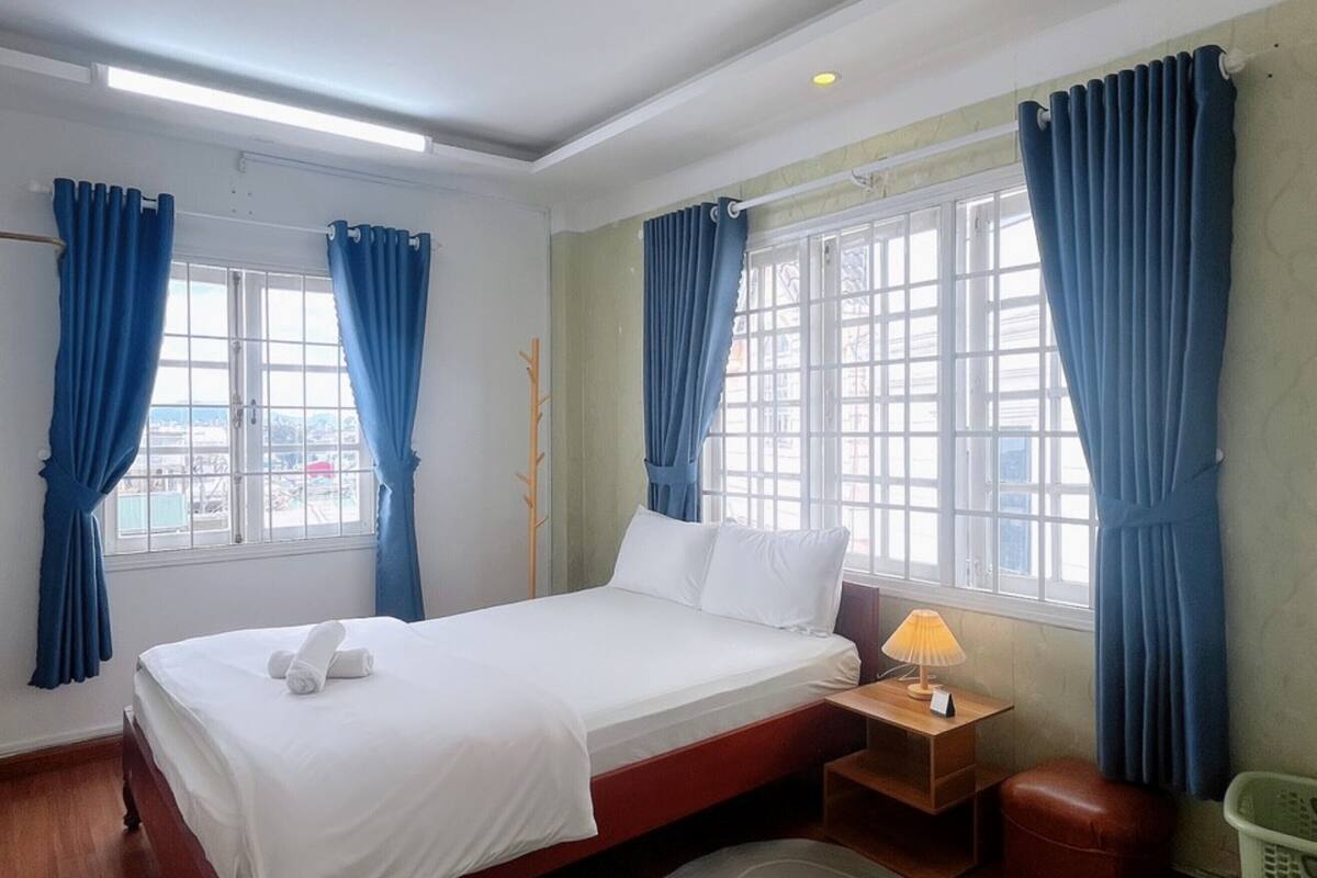 A hotel room with a bed, a nightstand with a lamp, a wooden floor, and a view through the window.