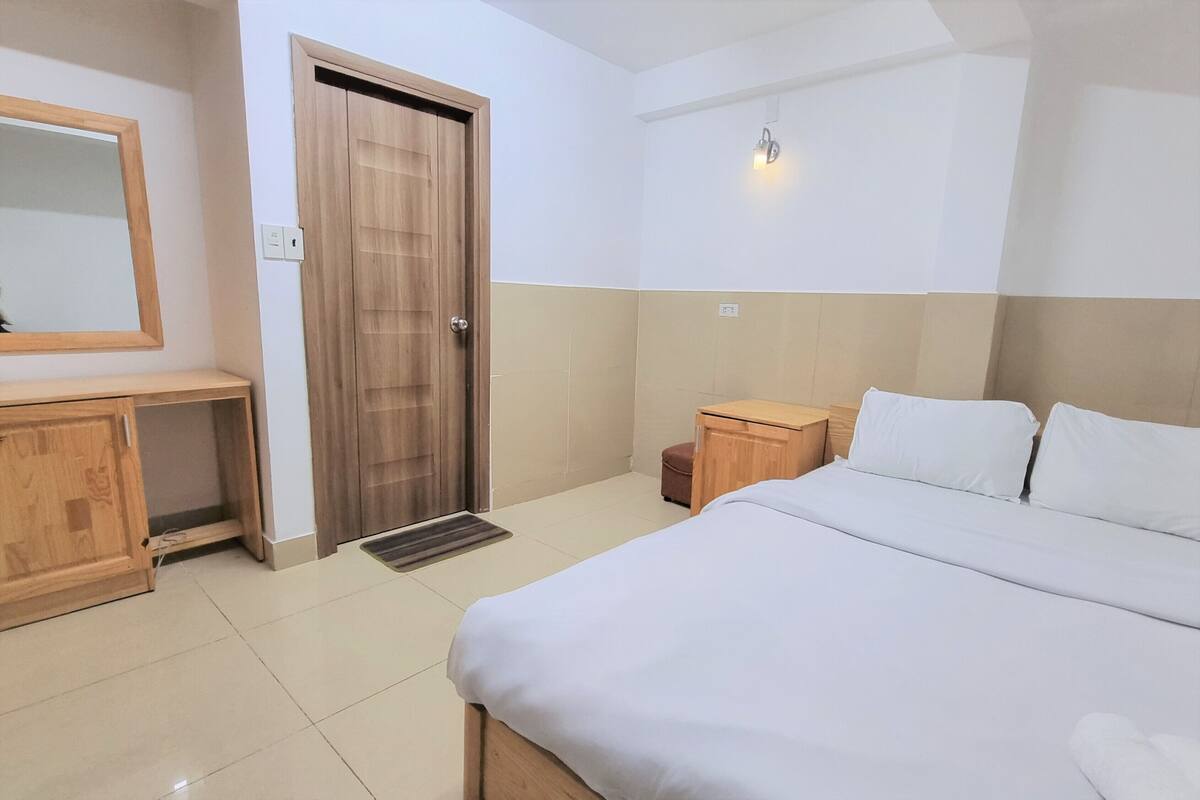 A hotel room with a bed, a wooden wardrobe, a small wooden nightstand, a mirror, and a door.