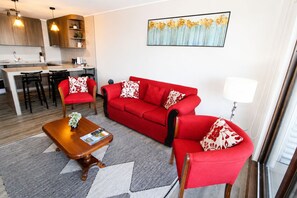 Ein modernes Wohnzimmer mit einem roten Sofa, passenden Sesseln, einem Holztisch und einer Küche im Hintergrund.