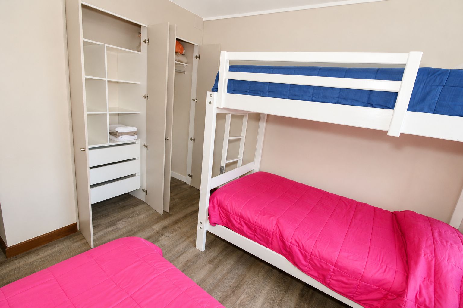 A bunk bed with a pink mattress and a white frame, a white wardrobe with multiple compartments, and a wooden floor.