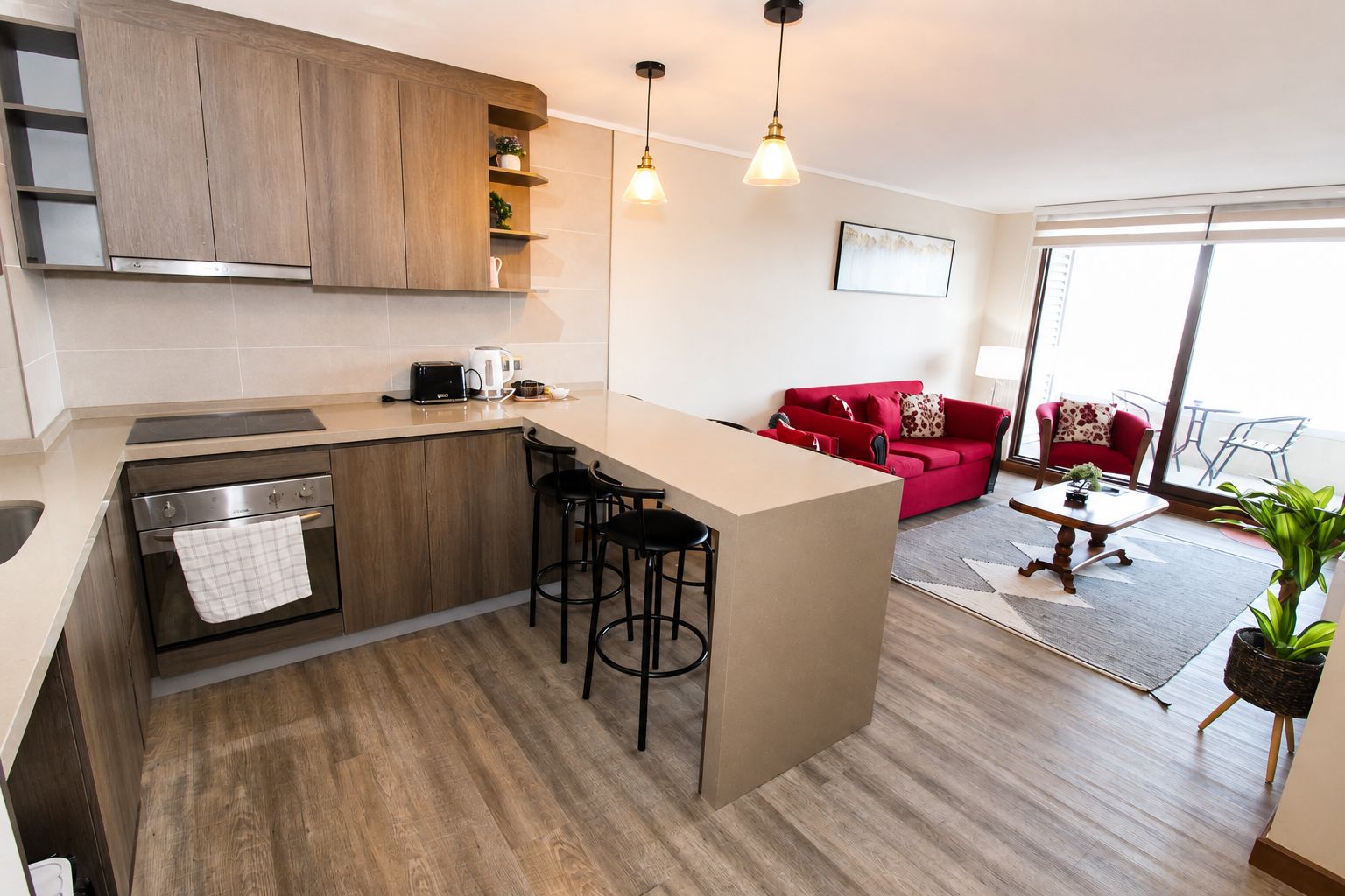 Een moderne keuken met een centraal eiland, barkrukken, en een rode bank in een woonruimte met uitzicht.