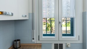 A kitchen with white cabinets, a stainless steel trash can, and a window with a blue frame and lace curtains. - HouSmart Cimabue 1 (Bologna)