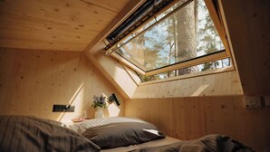 Un dormitorio acogedor con techo inclinado, tragaluz, cama con almohadas, mesita de noche con lámpara y una planta pequeña.