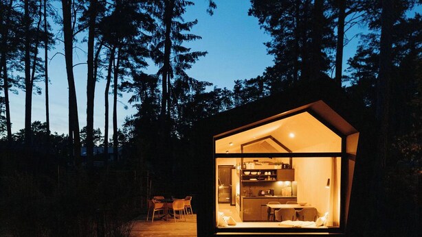 Una cabaña moderna e iluminada, con cocina y zona de comedor visibles a través de amplios ventanales, enclavada en un paisaje boscoso.