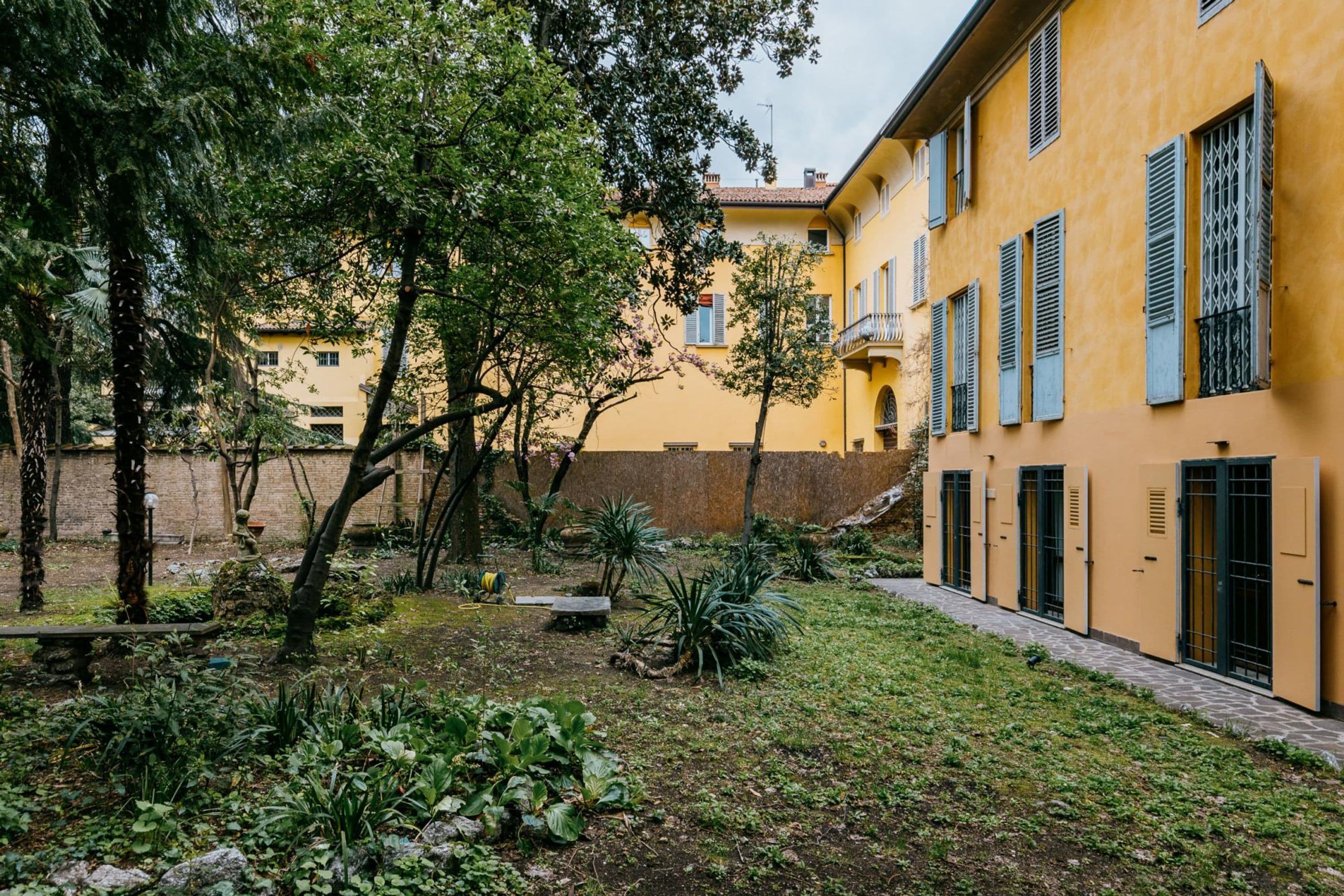 Une cour avec un bâtiment jaune, des arbres et un mur de pierre.
