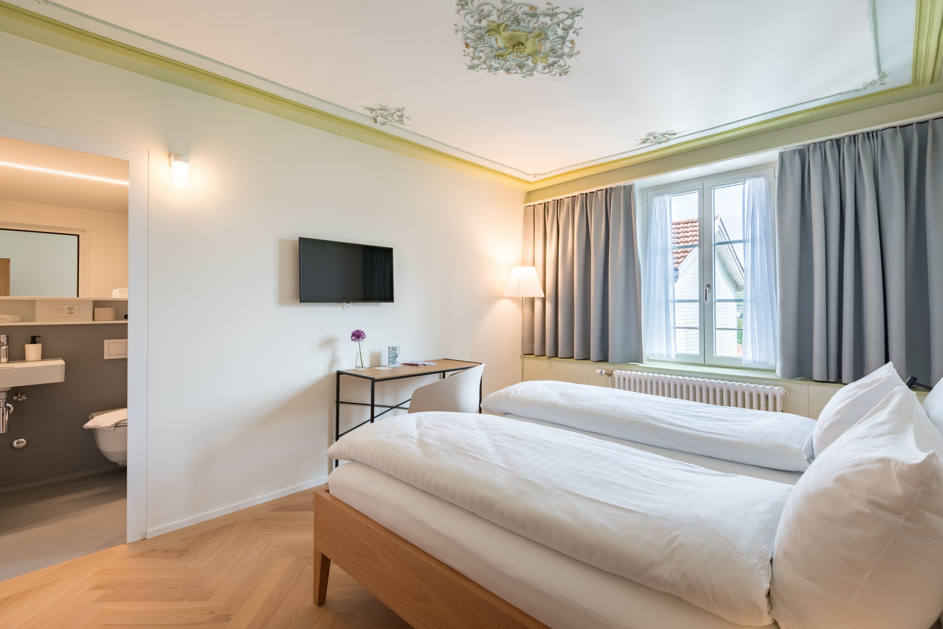 Una habitación de hotel con una cama de madera, un escritorio con silla, un televisor montado en la pared, una ventana con cortinas y un baño con lavamanos e inodoro.