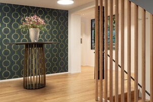 Un interior moderno con una pared estampada, suelo de madera, una mesa redonda con un florero y una escalera con barandillas de madera.