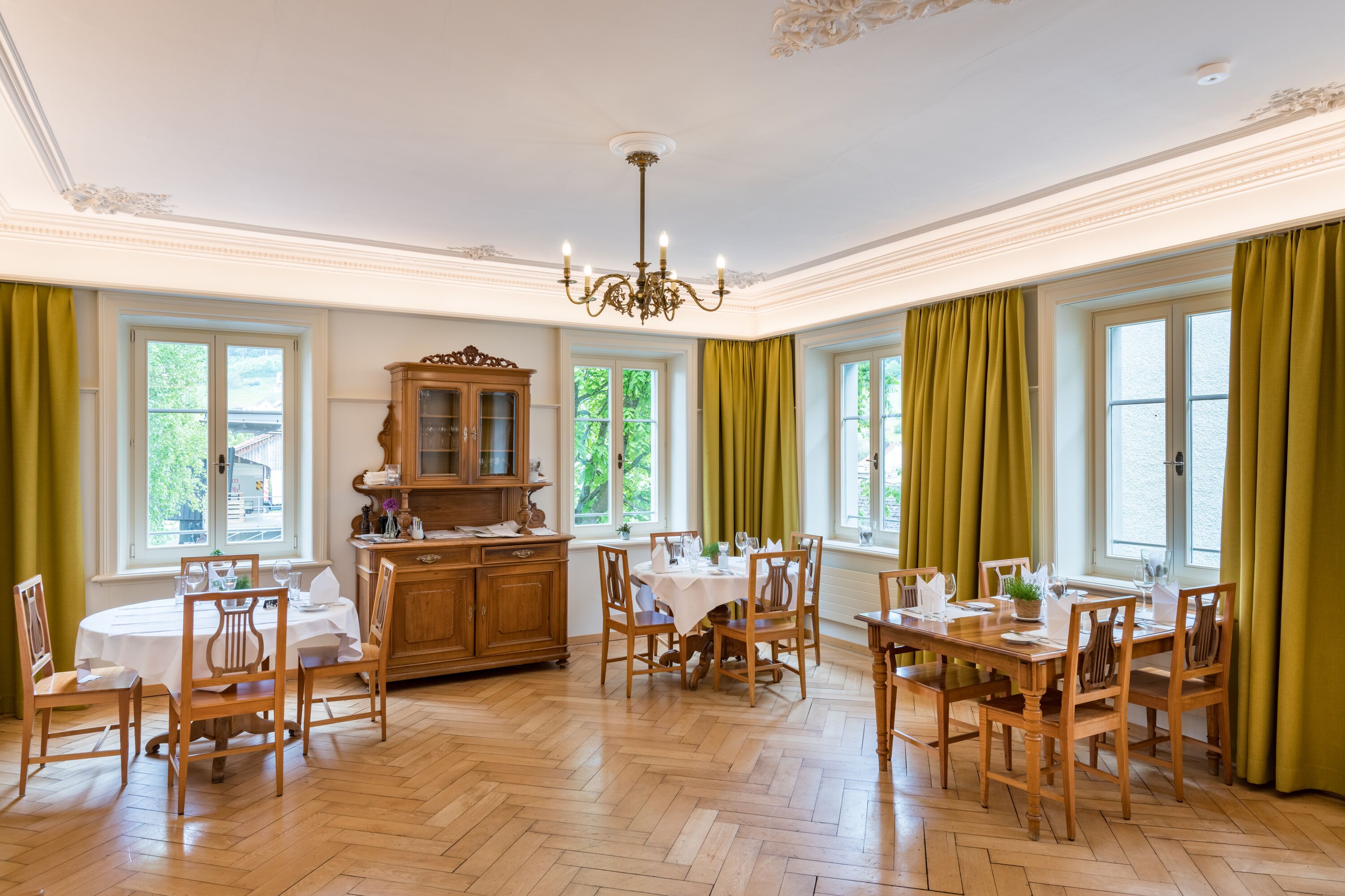 Ein Esszimmer mit Holzzmöbeln, einer Kristallleuchter und großen Fenstern mit Vorhängen.