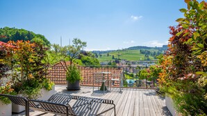 Een houten terras met een bankje en plantenpotten, met uitzicht op een groen landschap en een verder gelegen stad.