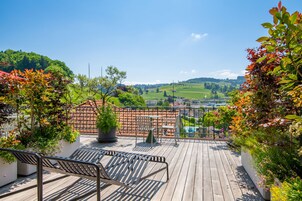 Una terraza de madera con un banco y plantas en macetas, con vistas a un paisaje verde y un pueblo lejano.