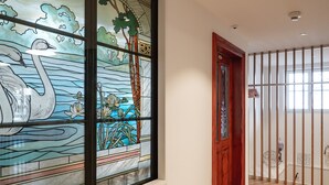 Een gang met een glas-in-loodraam waarop zwanen en waterlelies zijn afgebeeld, een houten deur, en uitzicht door een traliesraam.