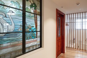 Un pasillo con un ventanal de vitral que muestra cisnes y nenúfares, una puerta de madera y una vista a través de una ventana con barrotes.