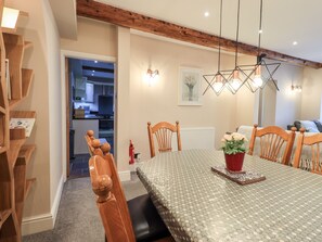 A dining area with a wooden table, chairs, and a chandelier. There is a kitchen visible in the background.