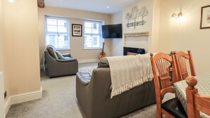 A living room with a sofa, a dining table with chairs, a fireplace, a TV mounted on the wall, and a window with blinds. - Bottle Tops (Windermere)