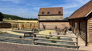 Un edificio rústico con fachada de piedra y techo inclinado, un patio con mobiliario y vistas al campo circundante.