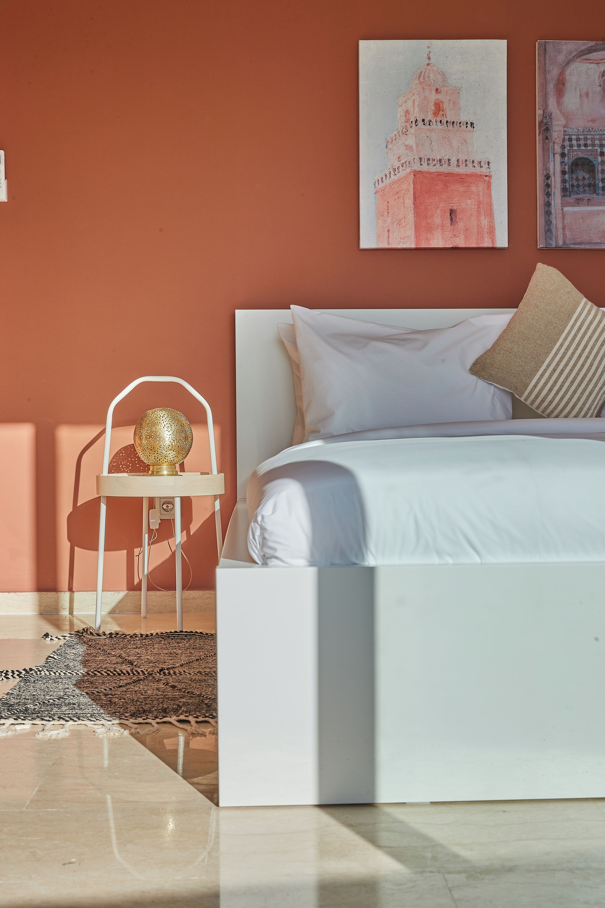 Een slaapkamer met een wit bed, een wit nachtkastje met een decoratieve lamp en een aan de muur hangend schilderij.