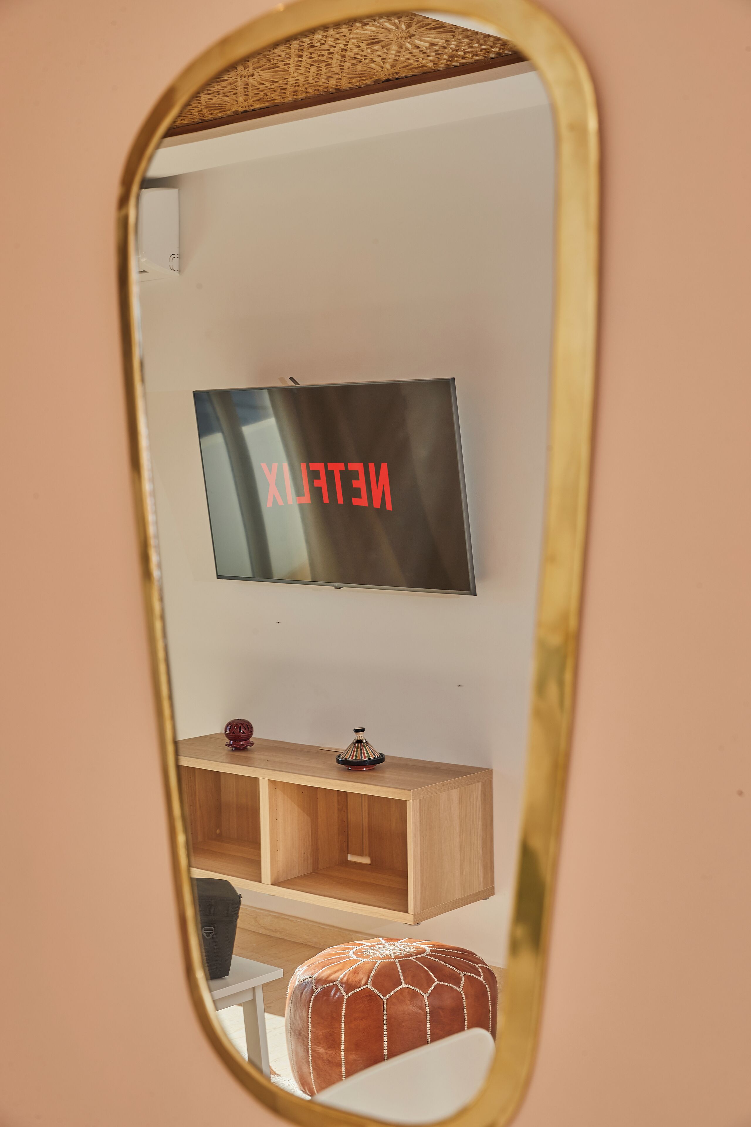 Un espejo que refleja una habitación con un televisor montado en la pared, un mueble de madera en la parte inferior y un otoman decorado.
