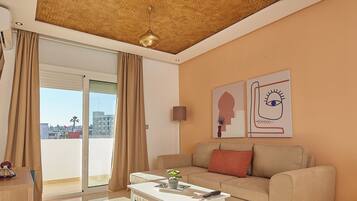 Una sala de estar moderna con un sofá, un reposapiés y una mesa de centro. Hay un ventanal con cortinas, un techo con diseño geométrico y una obra de arte en la pared.