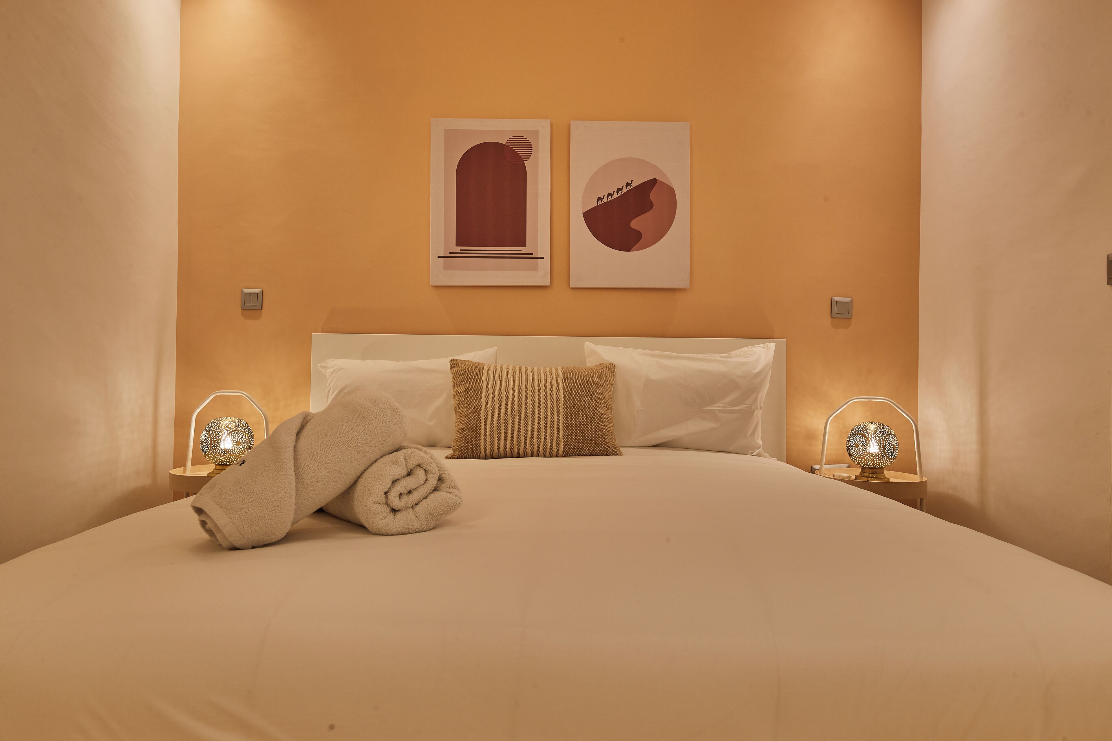 A neatly made bed with a rolled towel, two wall-mounted artworks, and bedside lamps.