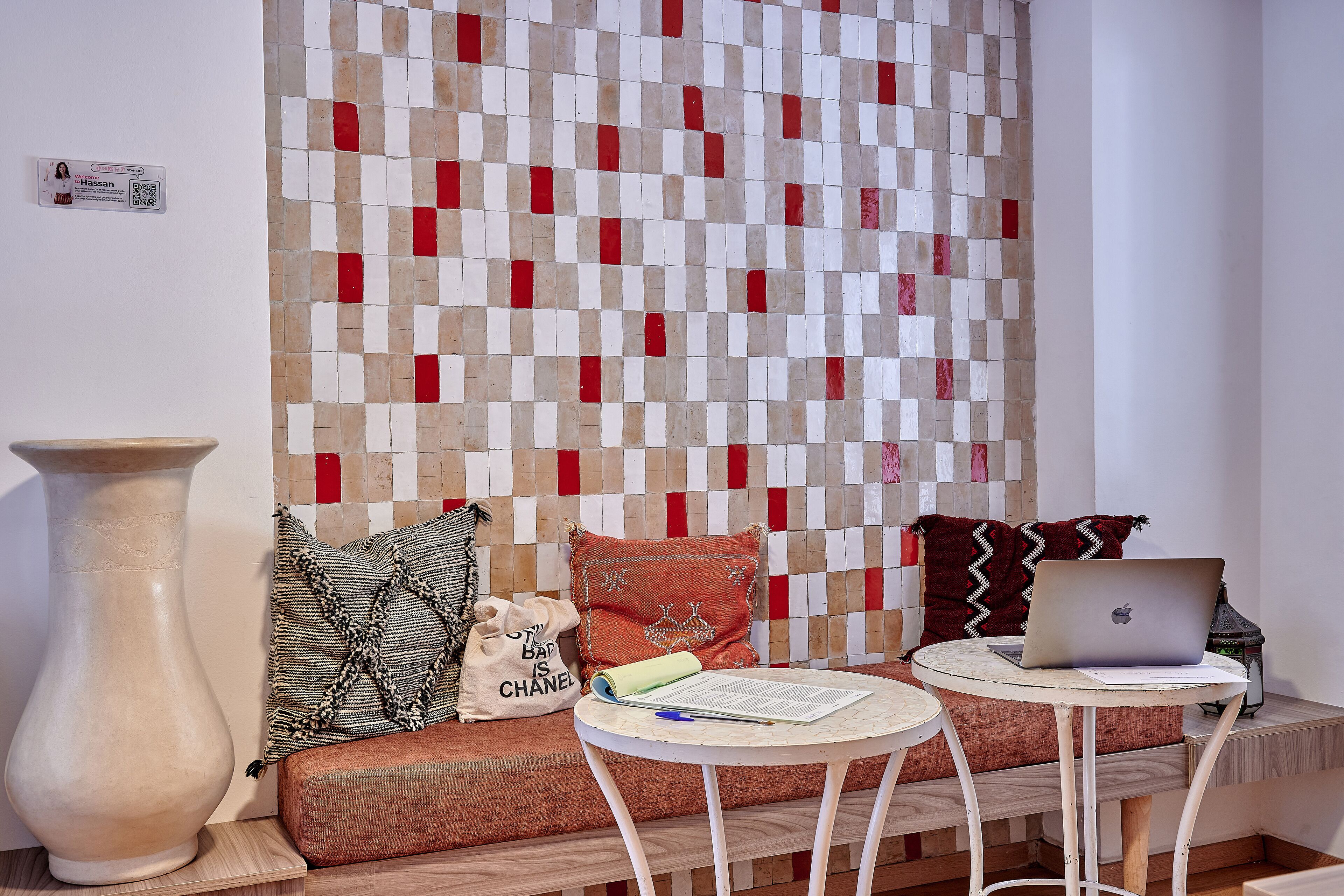 A room with a checkered wall, a sofa with cushions, a large vase, a small round table with a laptop, and a decorative lamp.