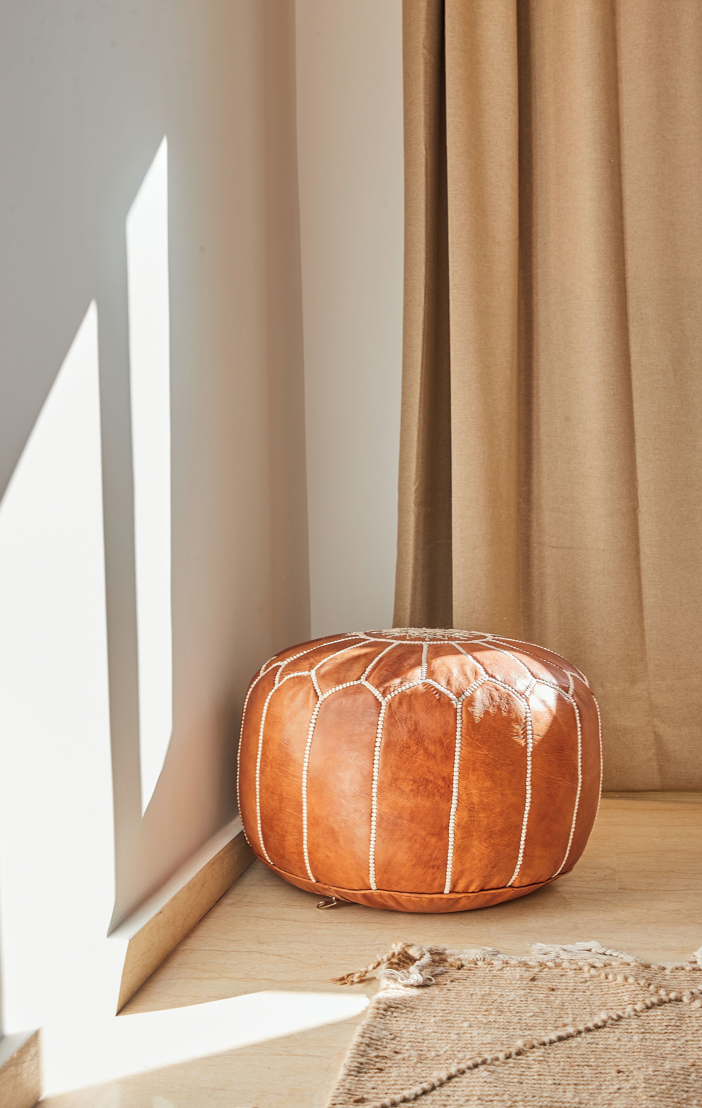 A brown leather pouf with white stitching, placed on a wooden ledge against a wall with beige curtains.