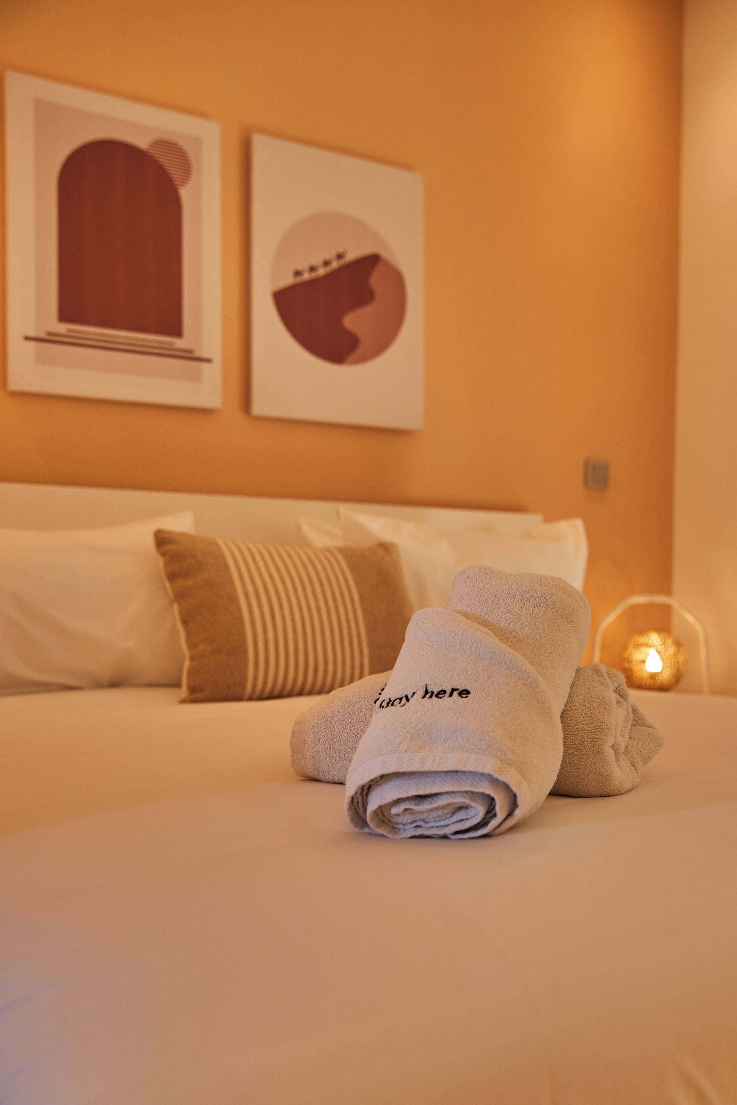 A neatly made bed with a rolled towel and a pillow on it. There is a wall with two framed pictures and a lit candle on a bedside table.