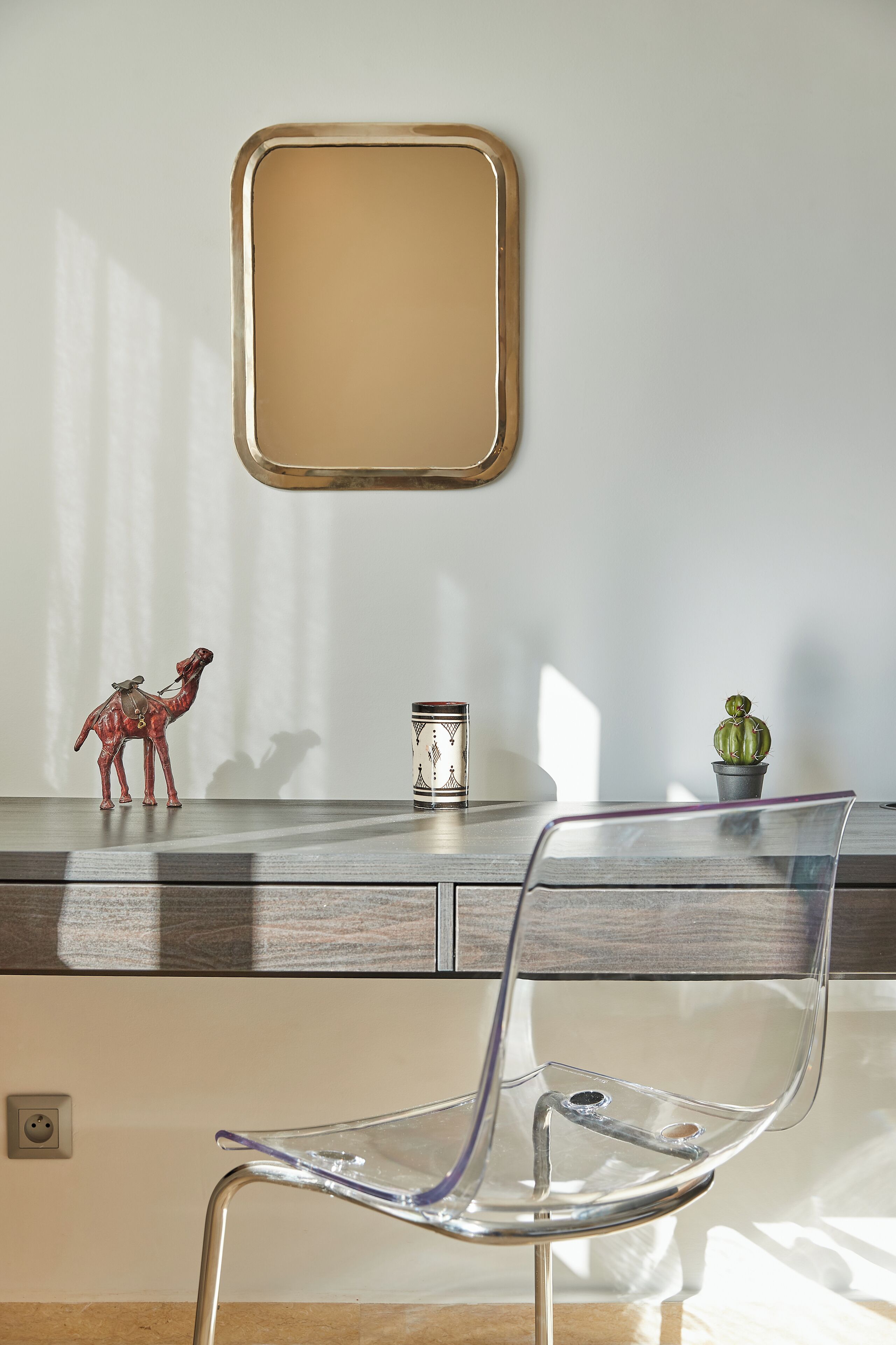 A clear glass chair in front of a desk with a small camel figurine, a cactus, and a decorative container.