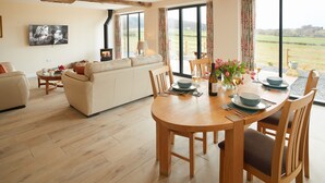 A spacious living area with a wooden dining table set for four, a sofa, and a television mounted on the wall. - A beautiful barn conversion sleeping four guests. (Acton Burnell, Shrewsbury)