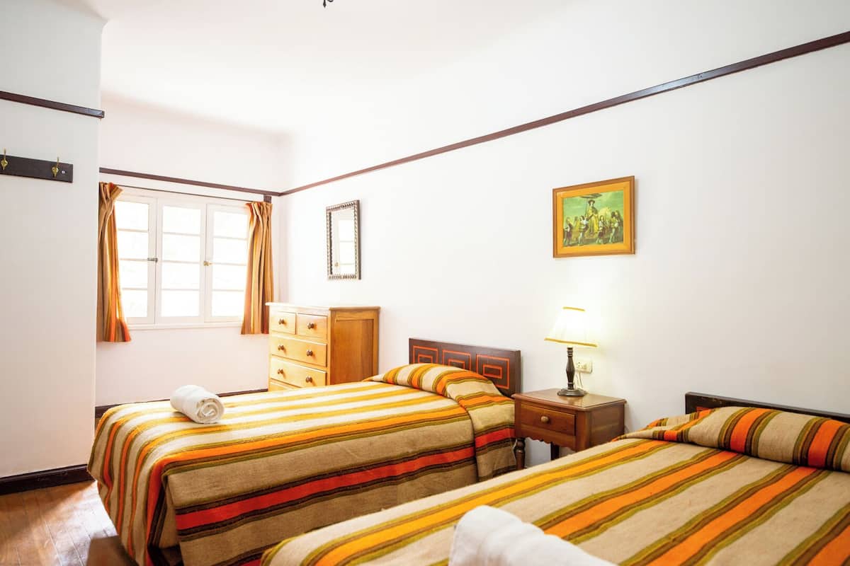 A hotel room with two beds, a wooden dresser, a nightstand with a lamp, a window with curtains, and a framed picture on the wall.