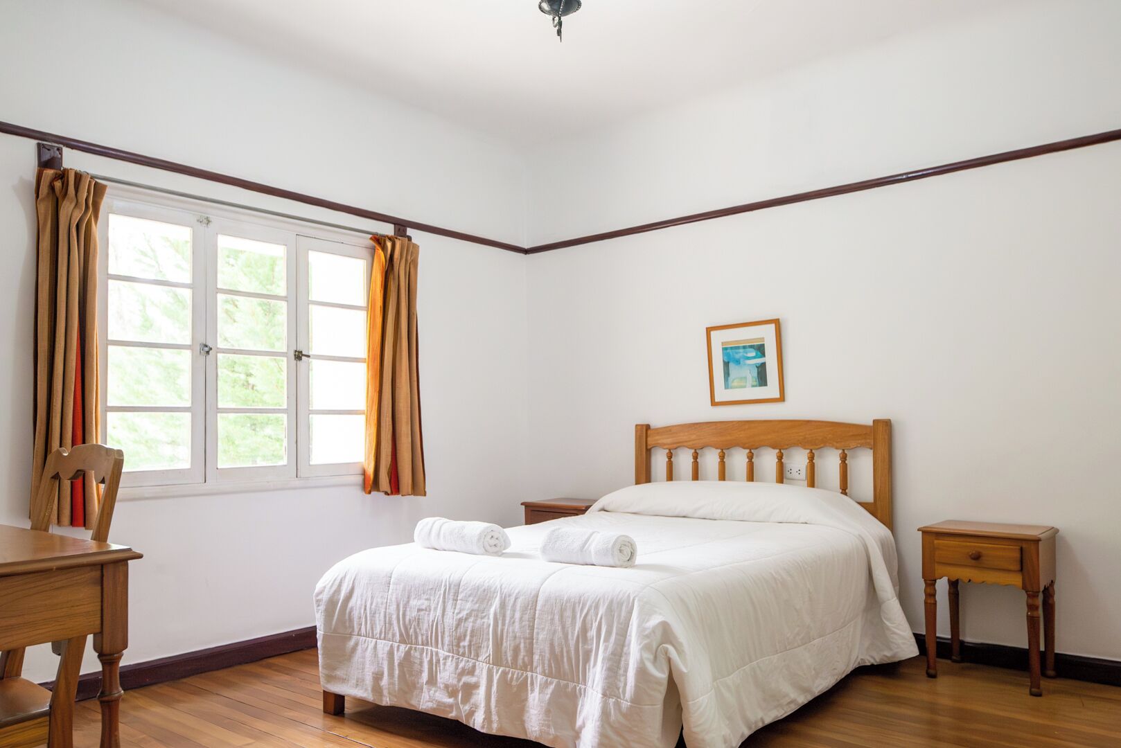 A bedroom with a bed, a chair, a nightstand, a window with curtains, and a framed picture on the wall.