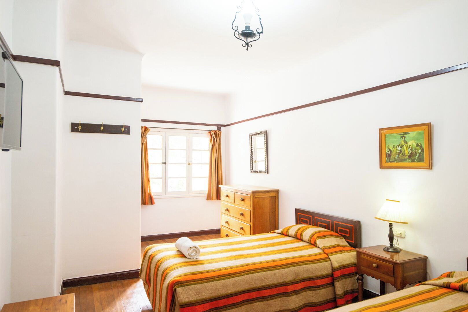 Une chambre d’hôtel comprenant un lit, une commode, une table de chevet, une lampe, une fenêtre avec des rideaux, un miroir et un tableau accroché au mur.