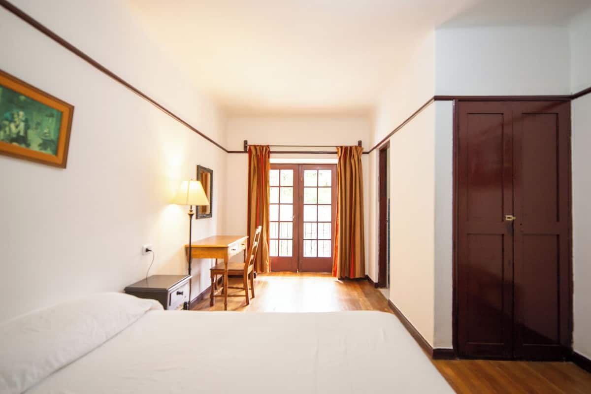 A bedroom with a bed, a desk, a chair, a lamp, a wardrobe, and a window with curtains.