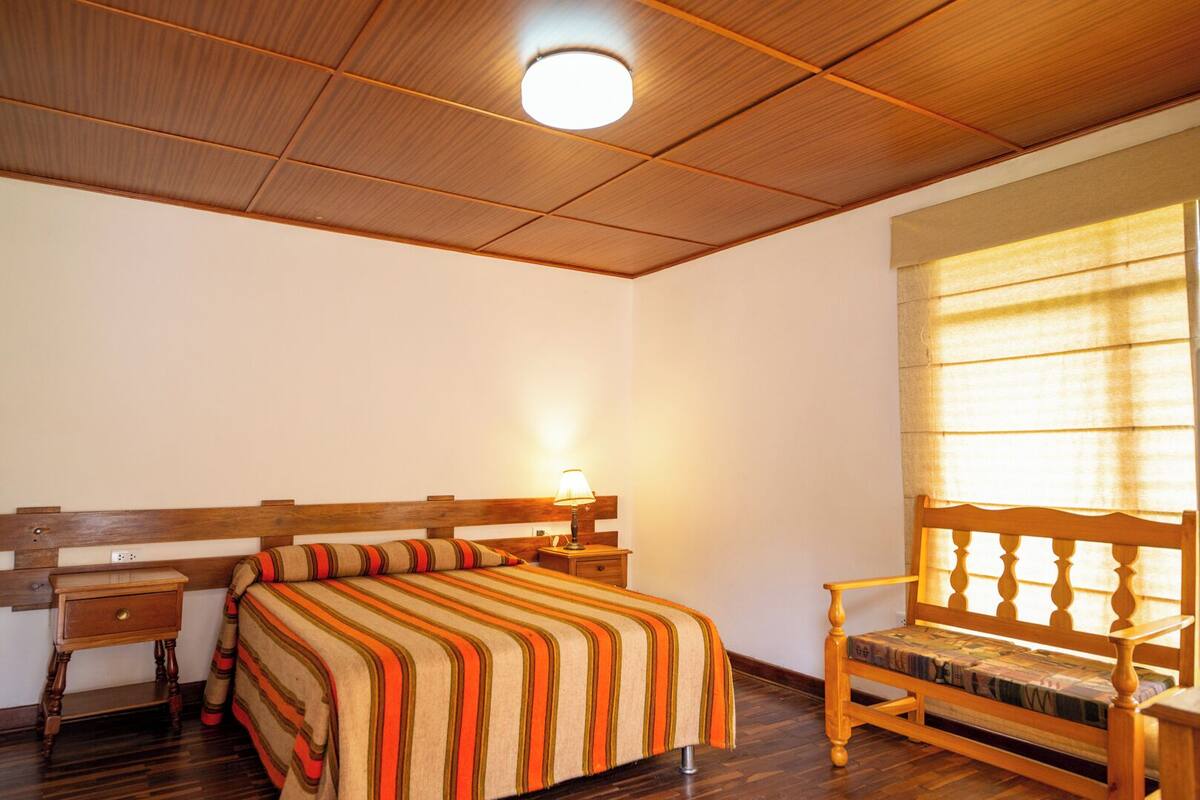 A bedroom with a wooden bed, a bedside table, a chair, and a window with blinds.